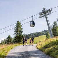 Drei Wanderer gehen einen Weg mit Seilbahn im Hintergrund.