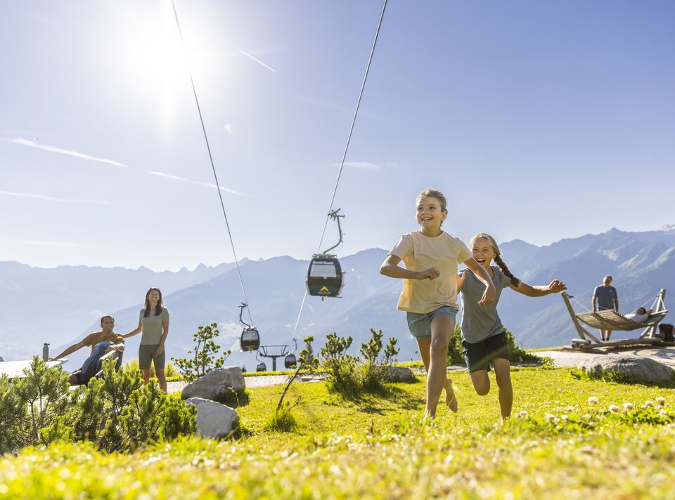 Two joyful girls run on a meadow, a cable car in the background.