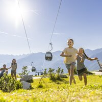 Zwei fröhliche Mädchen laufen auf einer Wiese, im Hintergrund eine Seilbahn.