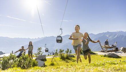Zwei fröhliche Mädchen laufen auf einer Wiese, im Hintergrund eine Seilbahn.