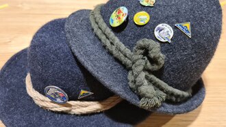Zwei dunkle Hüte mit verschiedenen Pins, auf Holzuntergrund.