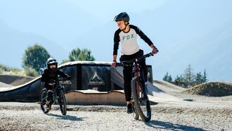 A boy and a child riding bicycles in a bike park surrounded by mountains.