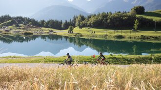 Zwei Radfahrer fahren am Ufer eines klaren Sees zwischen Bergen.