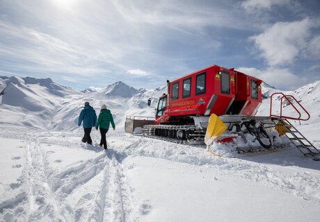 Two people walking in a snowy landscape beside a red snow vehicle.