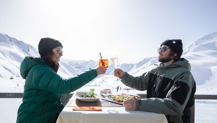 Ein Paar hebt Gläser auf einer Bergterrasse mit Blick auf die verschneiten Berge.