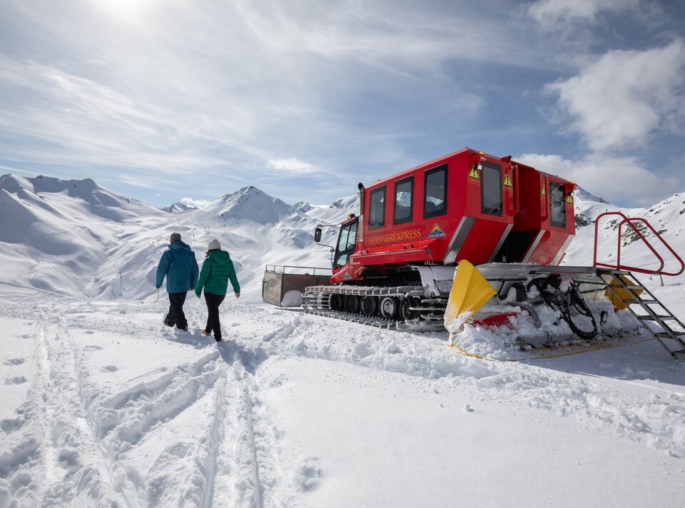 Zwei Personen spazieren in einer schneebedeckten Landschaft bei einem roten Schneefahrzeug.