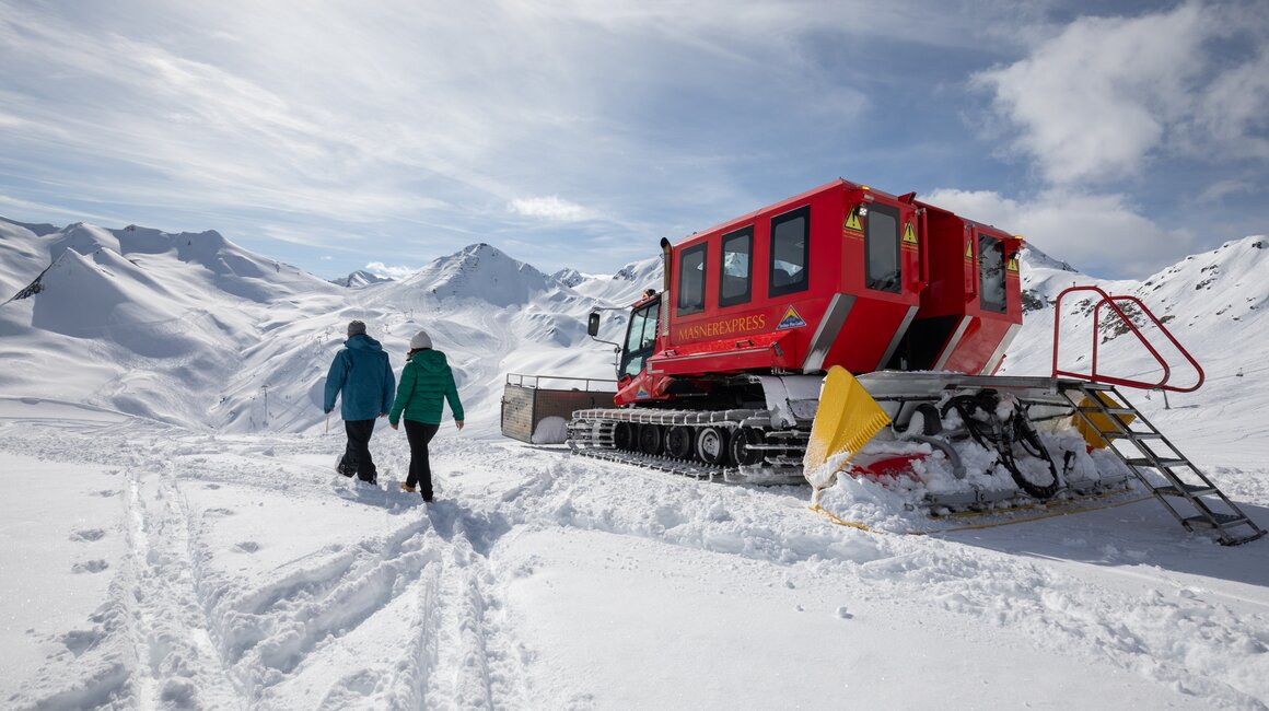 Zwei Personen spazieren in einer schneebedeckten Landschaft bei einem roten Schneefahrzeug.