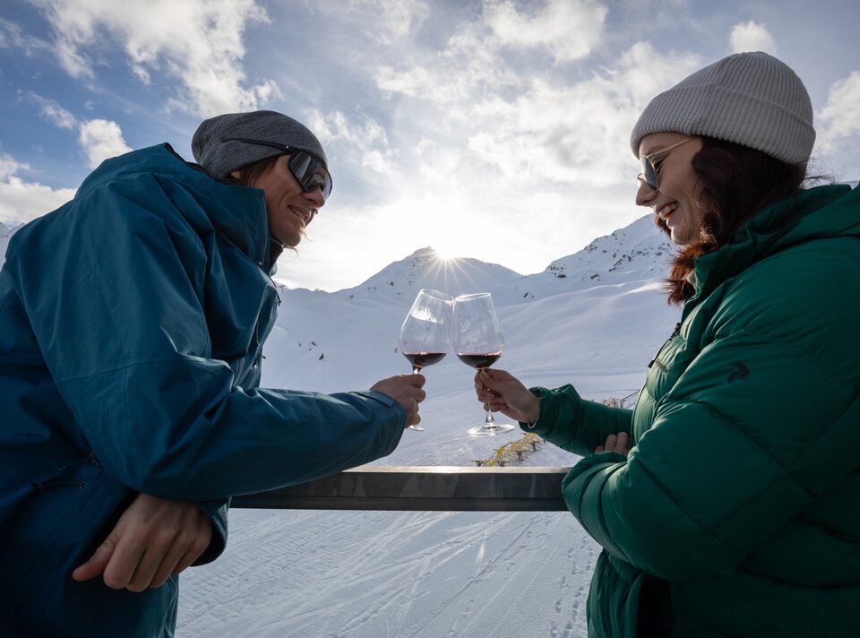 Zwei Menschen in Winterkleidung stoßen mit Rotwein auf der Schneepiste an.