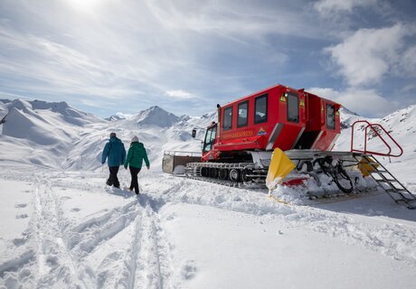 Two people walking in a snowy landscape beside a red snow vehicle.