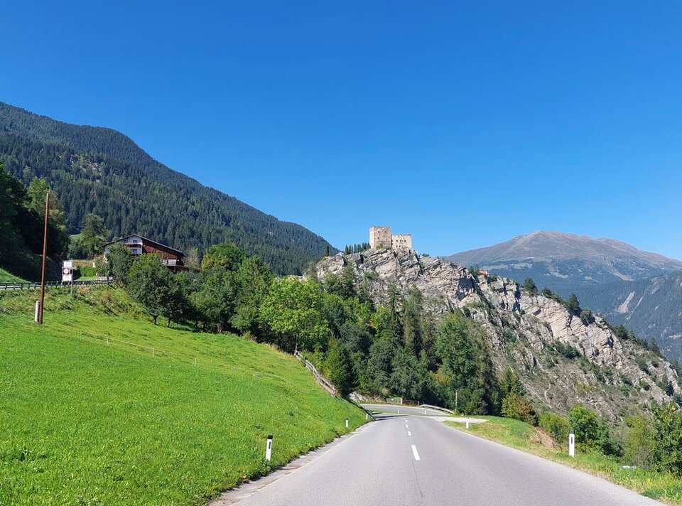 Eine Straße führt vorbei an einer Burg auf einem Berg, umgeben von grüner Natur und Bergen.