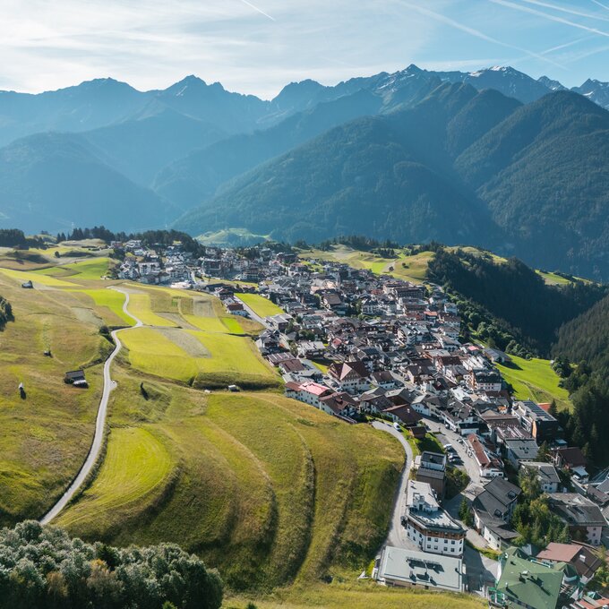 Luftaufnahme einer Bergstadt mit grünen Wiesen und Bergen im Hintergrund.