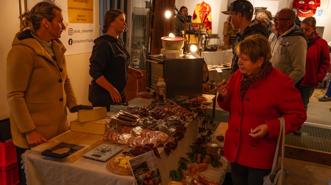Visitors explore a stall with cheese, sausage, and more at a market.