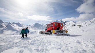 Zwei Personen wandern durch den Schnee nahe einem roten Pistenfahrzeug.