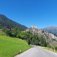 A road leads past a castle on a hill, surrounded by green nature and mountains.