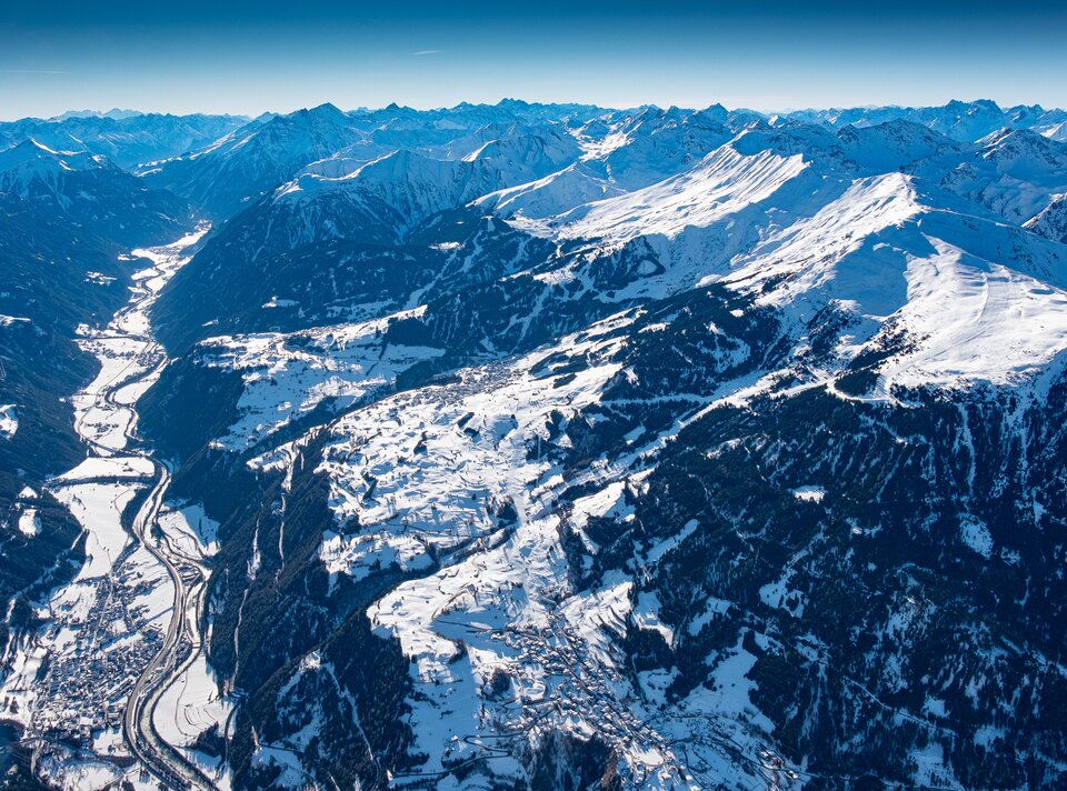 Luftaufnahme einer verschneiten Berglandschaft mit Tälern und Dörfern.