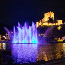 A blue illuminated fountain in front of a castle by the water at night.