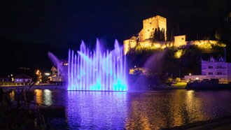 Ein blauer beleuchteter Brunnen vor einer Burg am Wasser während der Nacht.
