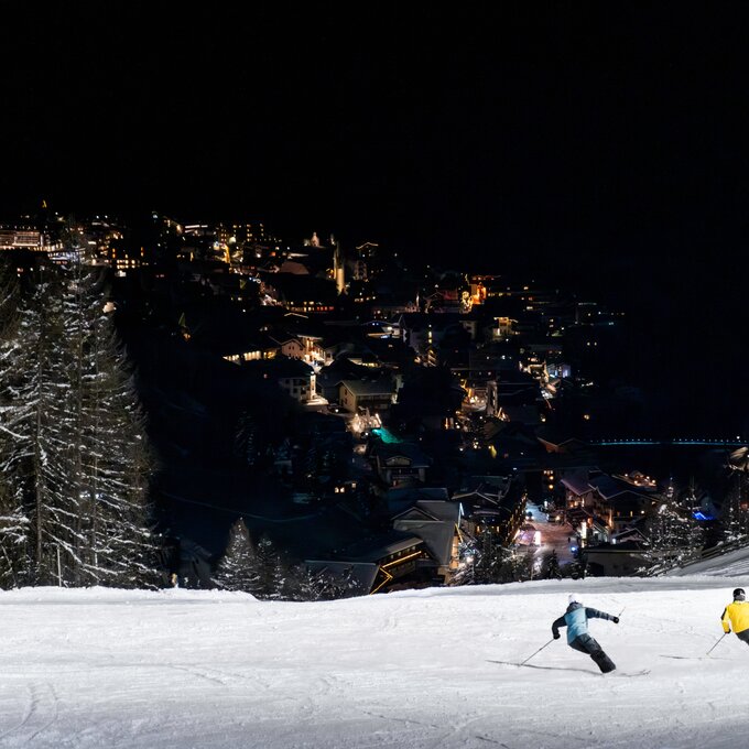 Twee skiërs glijden van een verlichte helling in de bergen, met een stad op de achtergrond.