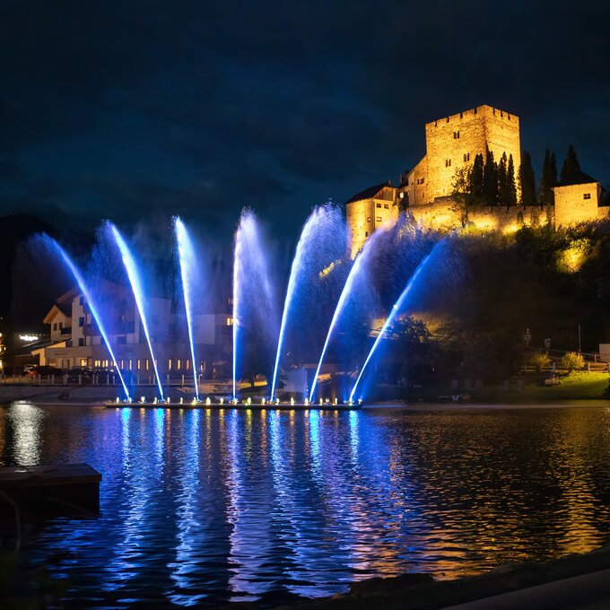 Licht-und-Wasser-Show am See mit einer Burgruine im Hintergrund.