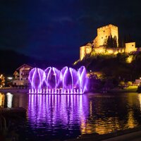 Abendstimmung mit beleuchteten Fontänen und einer Burg.