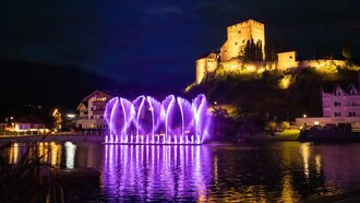 Abendstimmung mit beleuchteten Fontänen und einer Burg.