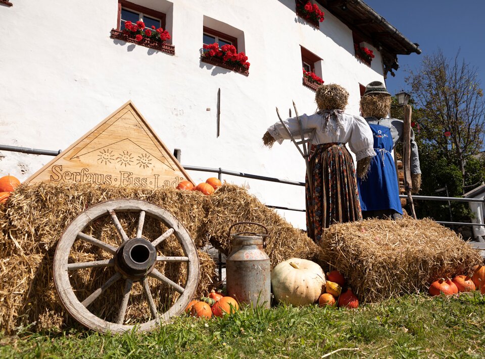 Zwei Strohpuppen vor einem Haus, umgeben von Kürbissen und Blumen.