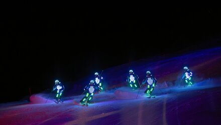 Multiple skiers in glowing outfits on a ski slope at night.