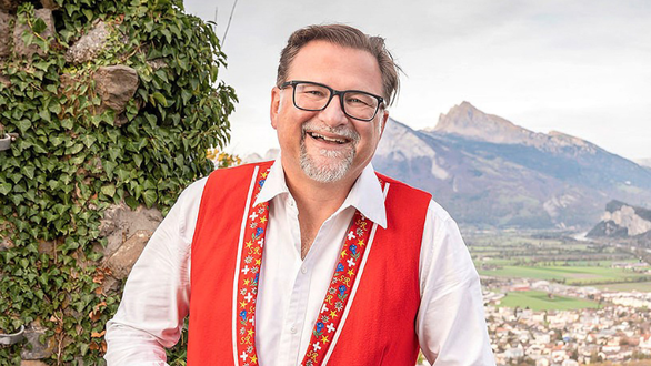 Freundlicher Mann mit Brille in rotem Weste vor Berglandschaft.