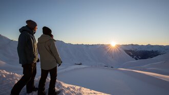 Two people standing in snow watching the sunrise.