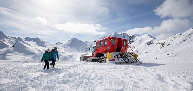 Zwei Personen wandern durch den Schnee nahe einem roten Pistenfahrzeug.