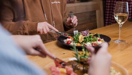 Zwei Personen speisen an einem Holztisch, mit Salat und Wein.