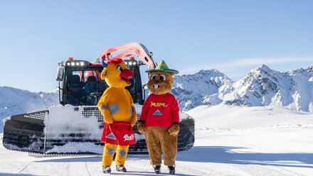 Two mascots stand in front of a snow groomer in the snow.
