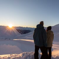 A couple admires the sunrise over snow-covered mountains.