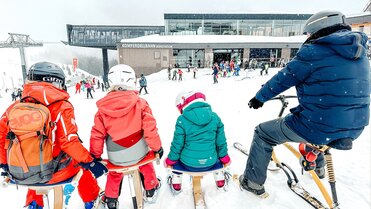 Familie sitzt im Schnee, blickt auf Skigebiet und Restaurant.