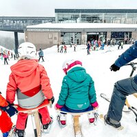 Familie sitzt im Schnee, blickt auf Skigebiet und Restaurant.