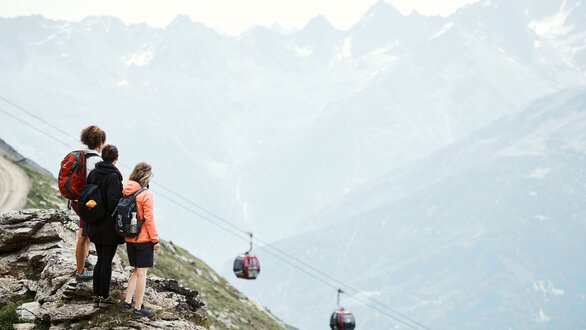 Drei Wanderer genießen die Aussicht auf Berge und Seilbahn.