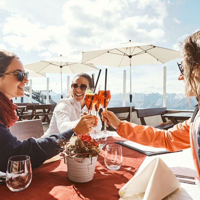 Drei Frauen stoßen mit Drinks auf der Terrasse eines Bergrestaurants an.
