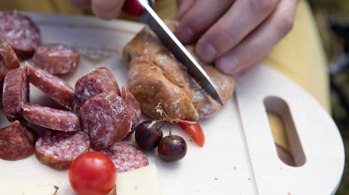 Eine Hand schneidet frisches Brot neben Salami, Käse und Trauben.