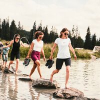 Drei Mädchen balancieren über Steine in einem Gewässer.