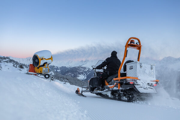 Ein Schneemobil bereitet die Piste vor, während ein Schneekanone im Hintergrund arbeitet.