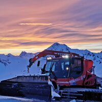 Eine Pistenraupe steht im Schnee unter einem farbenfrohen Sonnenuntergang.