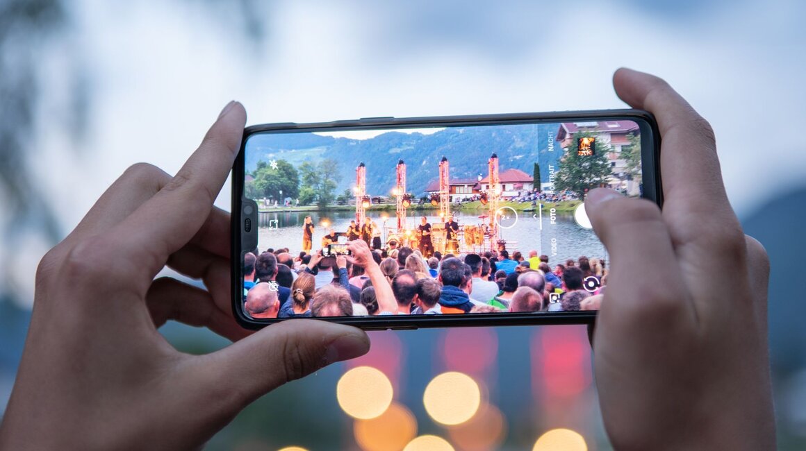 Eine Person filmt eine Show mit ihrem Handy am See, umgeben von Publikum.