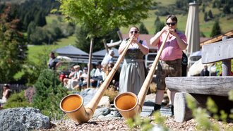 Zwei Musiker spielen Alphorn in einer malerischen Alpenlandschaft.