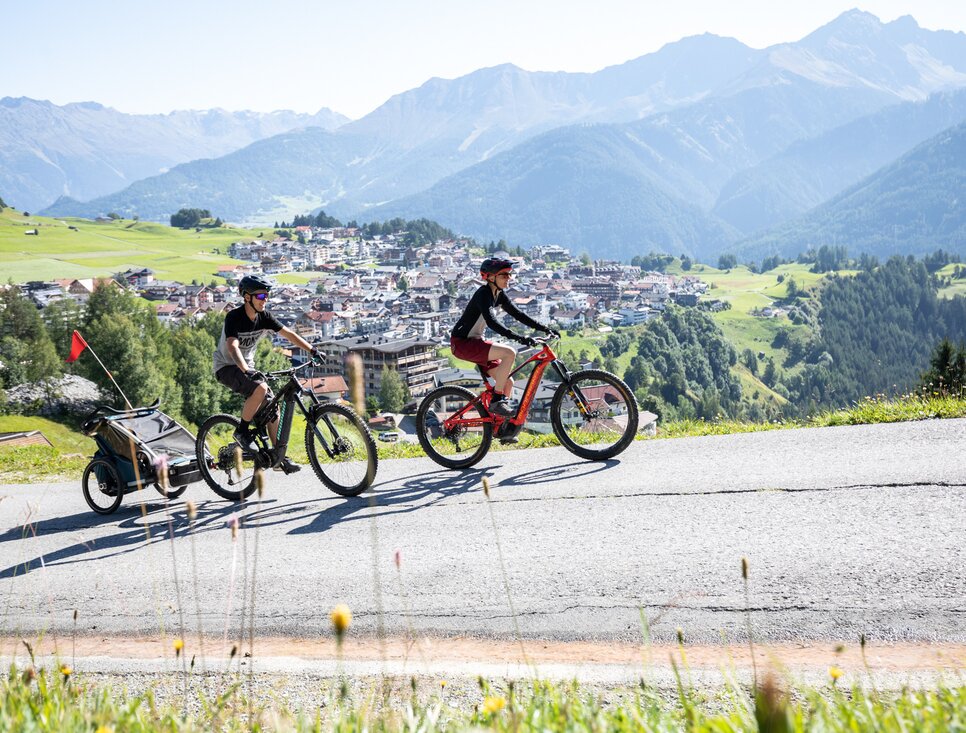 Zwei Radfahrer fahren eine Bergstraße mit schöner Landschaft und Stadtblick.