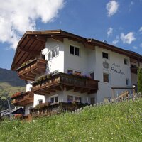 Blumengeschmücktes Apart Stecher mit Balkonen aus Holz in der sommerlichen Landschaft von Serfaus. Die Serfauser Berge mit den grünen Wiesen im Hintergrund erstrahlen in der warmen Sommersonne.