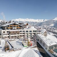 Eine Winterlandschaft mit verschneiten Häusern und Bergen im Hintergrund. Es gibt Spielgeräte im Vordergrund und eine ruhige Atmosphäre.